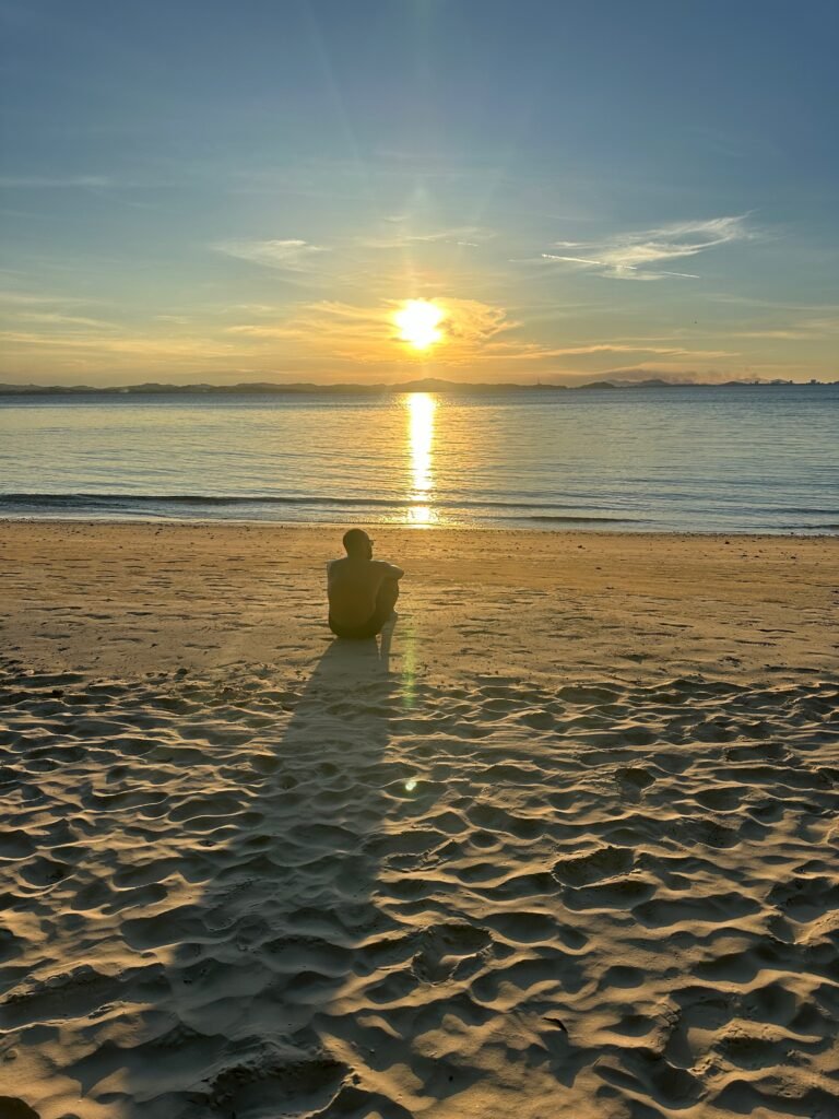 A man is sitting by the beach watching the sunset