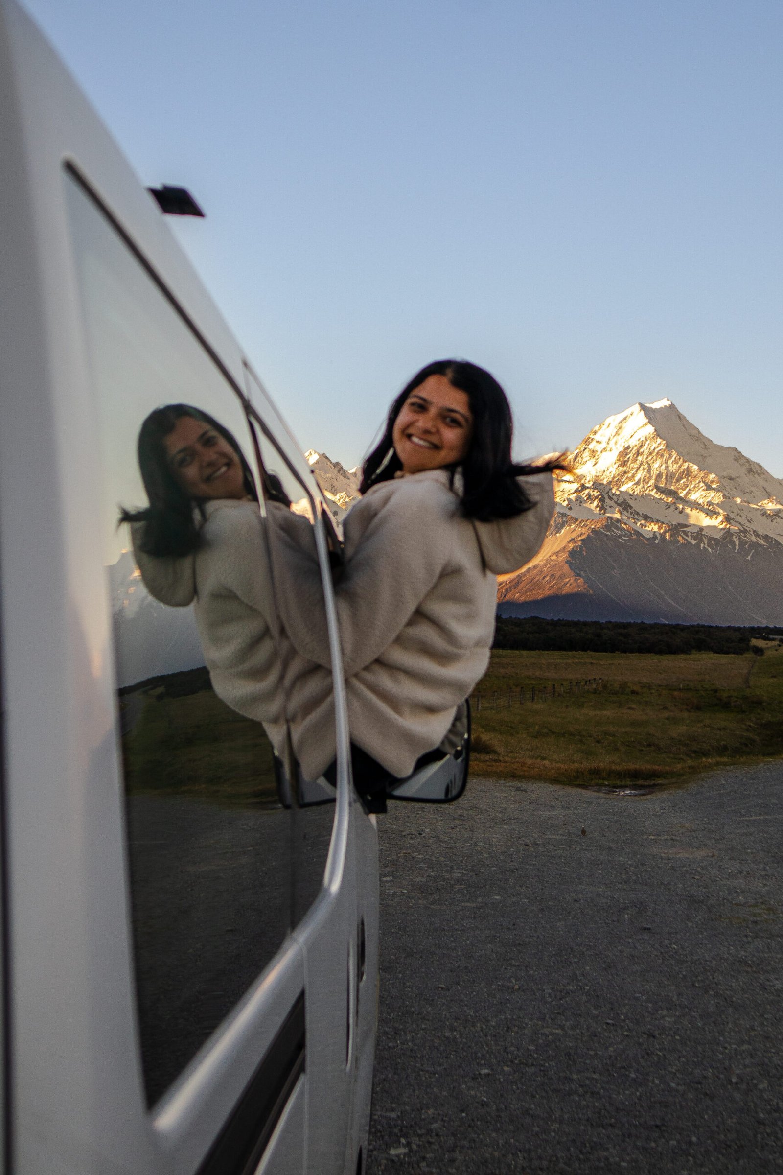 Woman sitting on campervan window with Mount Cook in the background, photographed from outside the van