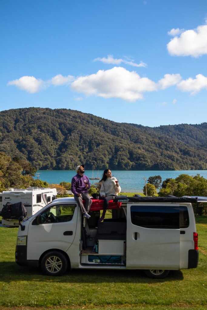 Couple sitting on top of a campervan enjoying a scenic New Zealand landscape. The post discuss New Zealand Travel costs