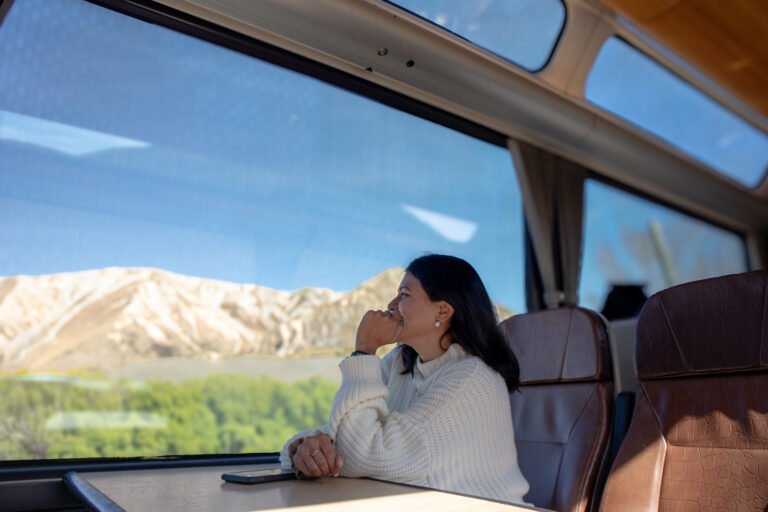 Woman seated inside the TranzAlpine train watching the Southern Alps through panoramic windows in New Zealand