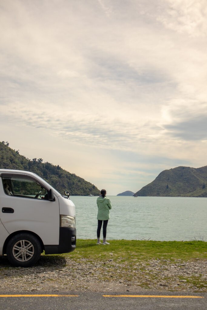 Person standing by a lake with a campervan parked nearby, mountains in the background