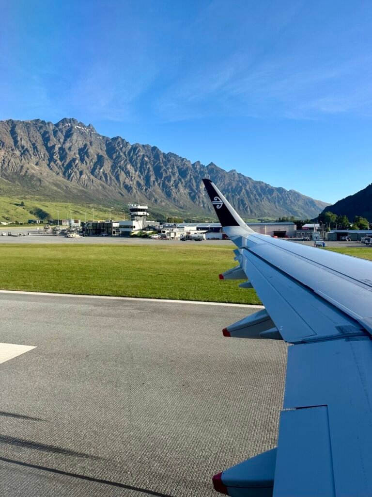 View from airplane window showing Air New Zealand wing over Queenstown Airport with The Remarkables mountain range in the background