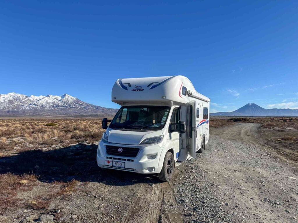 White Jayco motorhome parked on a gravel track on the Tongariro volcanic plateau with snow-capped volcanoes in the background