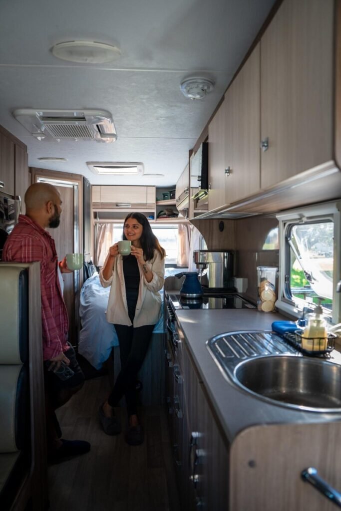 Couple holding mugs and chatting inside the kitchen of a motorhome, showing the sink, countertop and living space