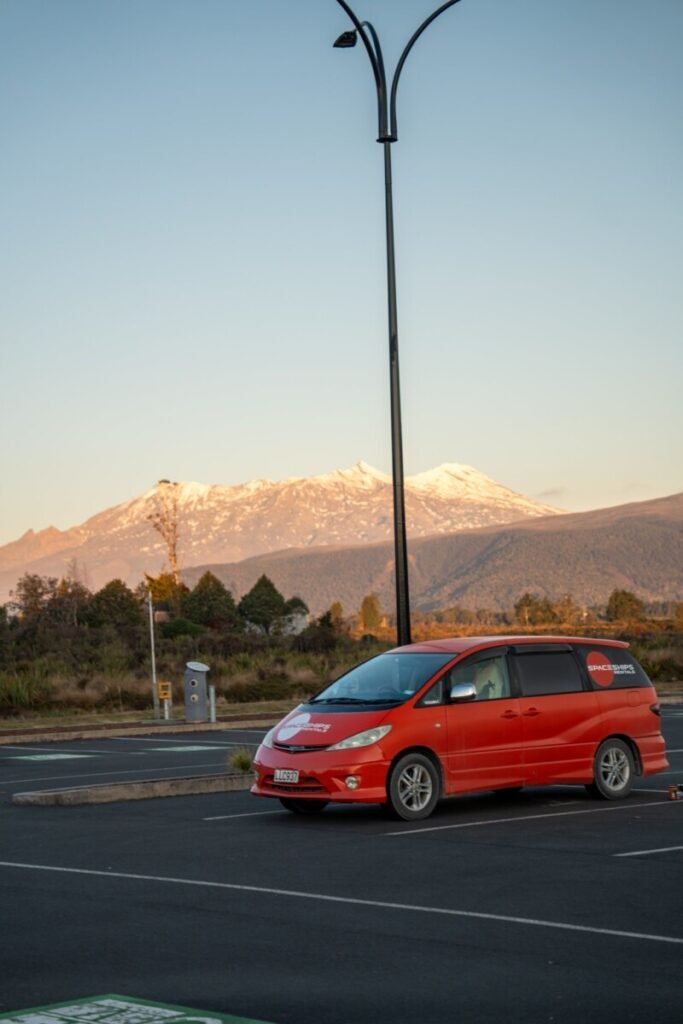 Red rental campervan parked in a car park with a snow-capped New Zealand volcano glowing in golden hour light
