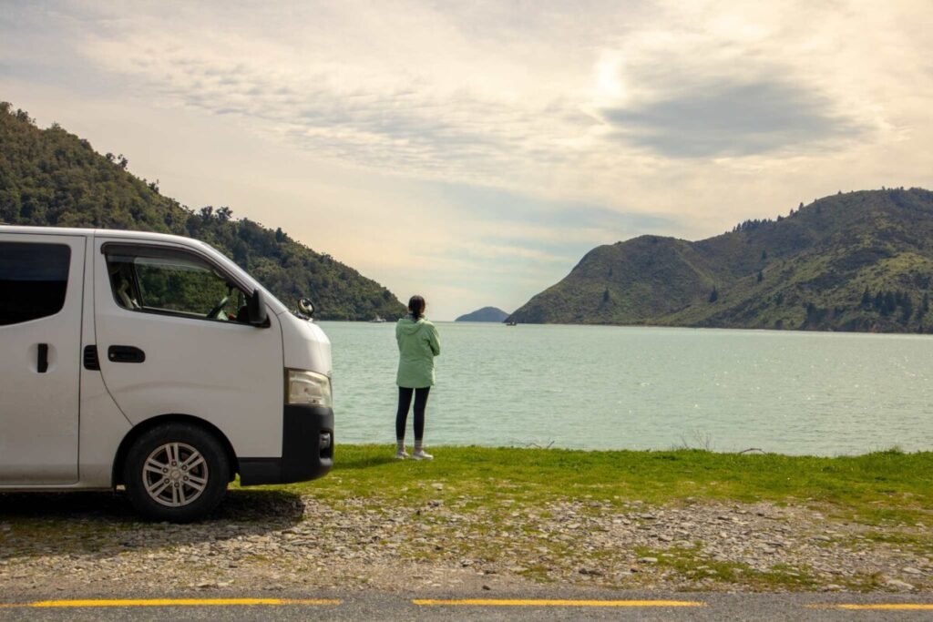 Woman standing beside a white campervan New Zealand overlooking a turquoise bay surrounded by green hills