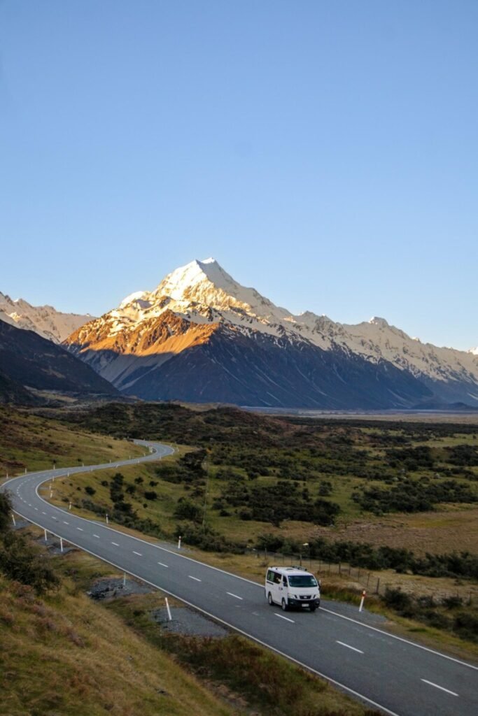White campervan driving along an open road toward snow-capped Aoraki Mount Cook glowing in golden sunrise light in New Zealand