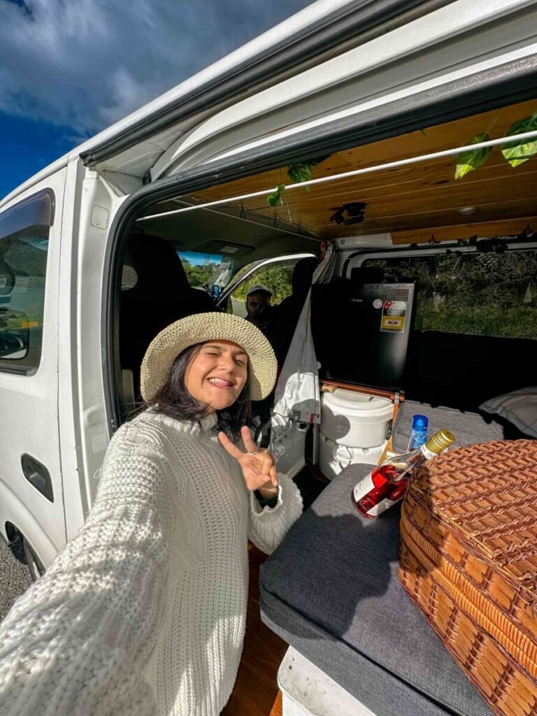 Woman in a straw hat smiling and making a peace sign at the open back door of a campervan with wine and a picnic basket inside