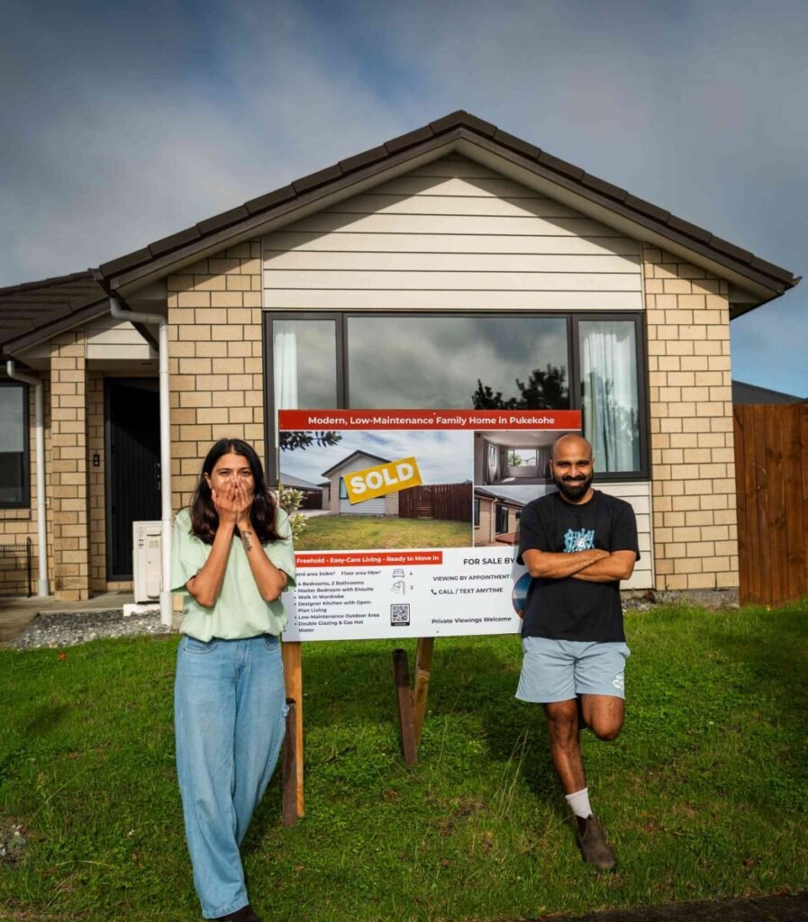 Couple standing in front of their sold house with a SOLD sign, about to start a campervan New Zealand road trip