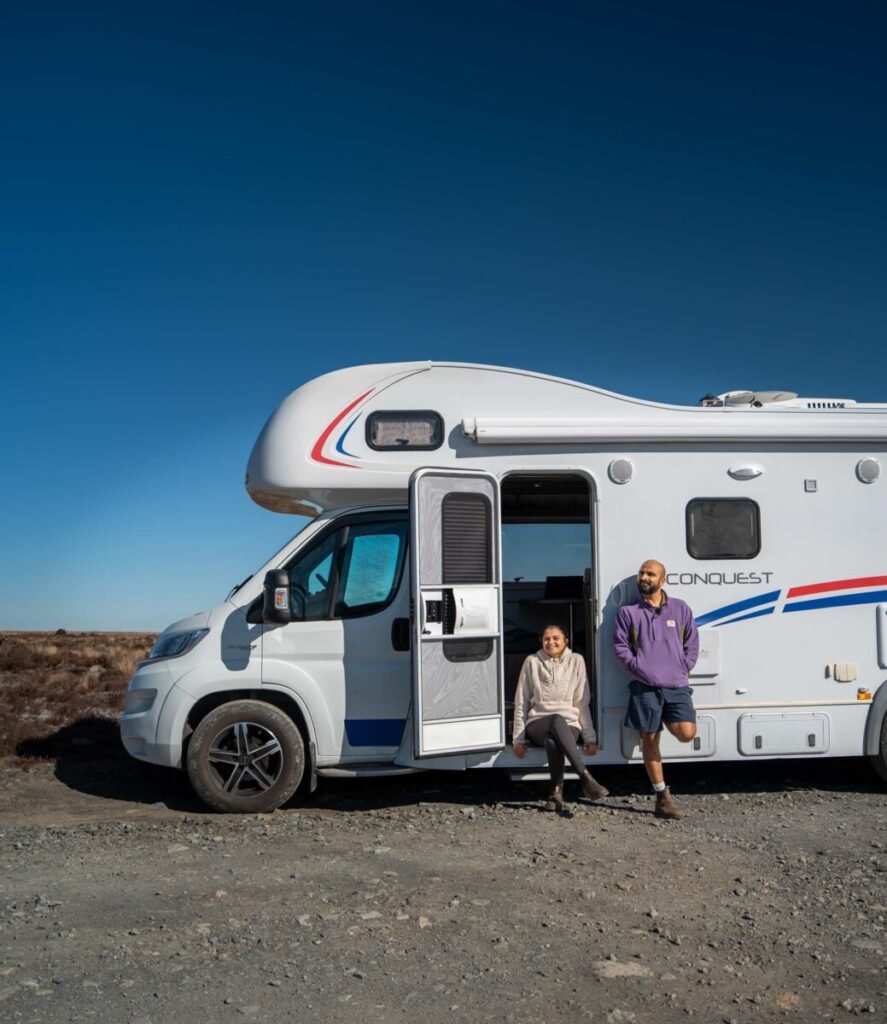 Couple posing at the open door of a white Conquest motorhome in a rocky New Zealand landscape under a clear blue sky