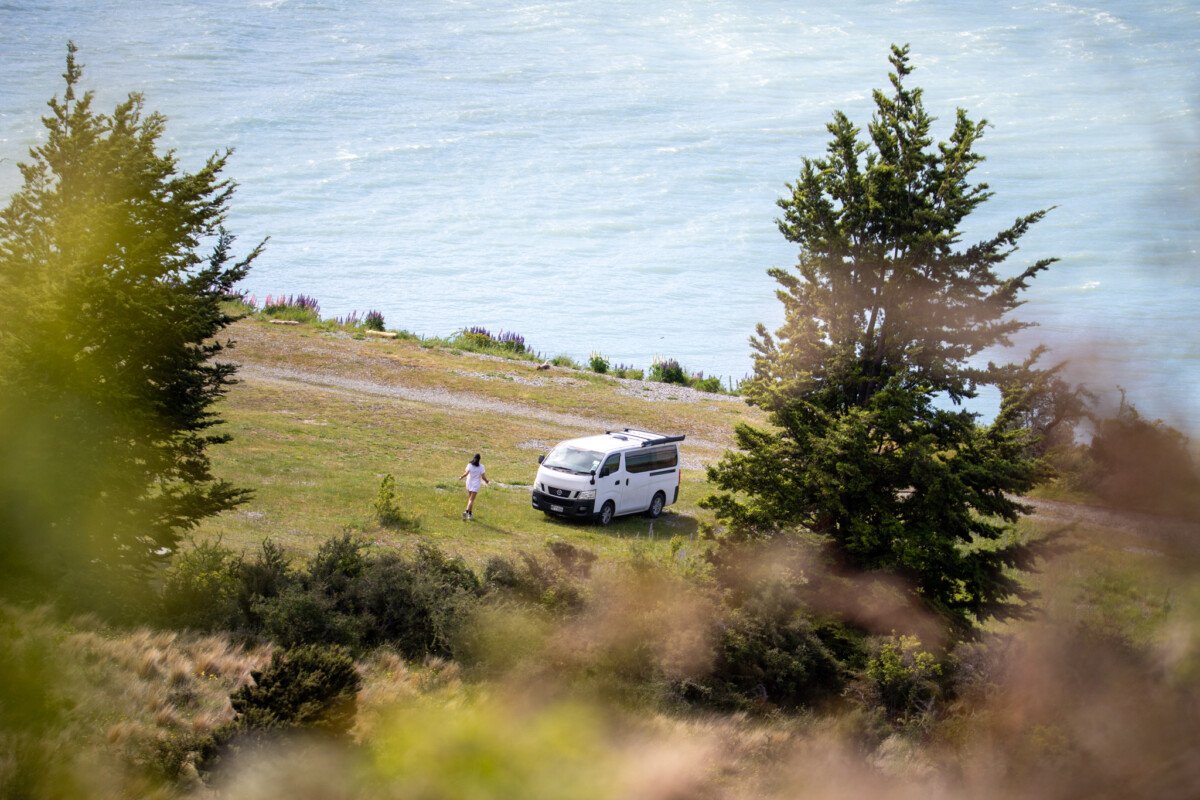 Woman walking towards a white campervan parked on a grassy coastal hillside with the ocean in the background while freedom camping in New Zealand