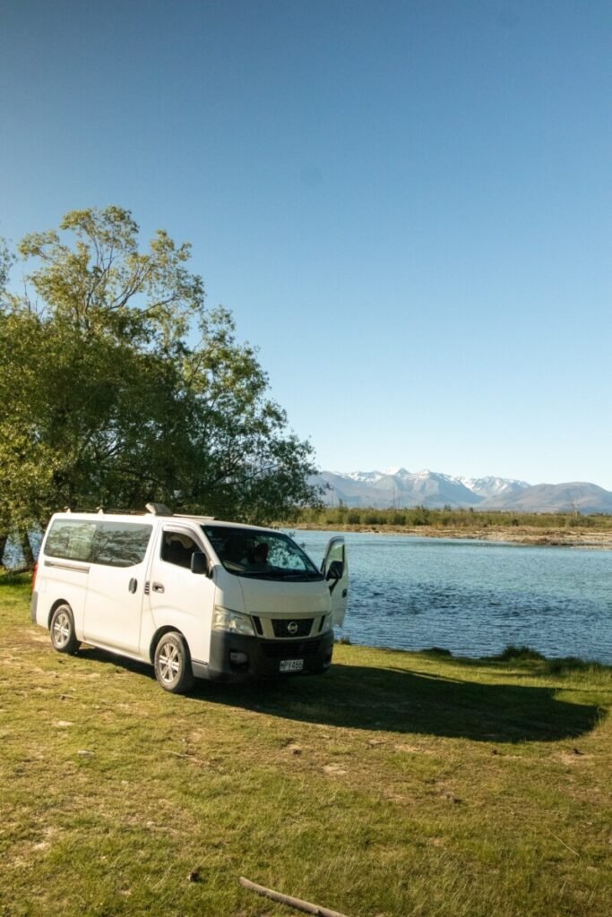 White campervan Wander Bee parked beside a blue glacial river with the Southern Alps in the background at a DOC campsite New Zealand