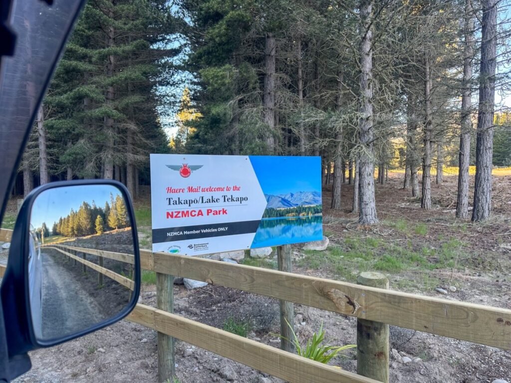 Welcome sign for Takapo Lake Tekapo NZMCA Park visible from the van wing mirror with pine trees in the background
