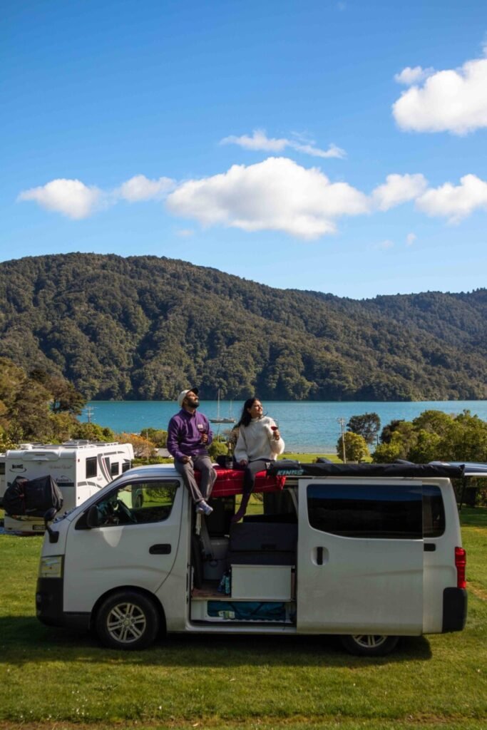 Couple sitting on top of their campervan at Momorangi Bay DOC campsite with turquoise Marlborough Sounds and forested hills in the background New Zealand