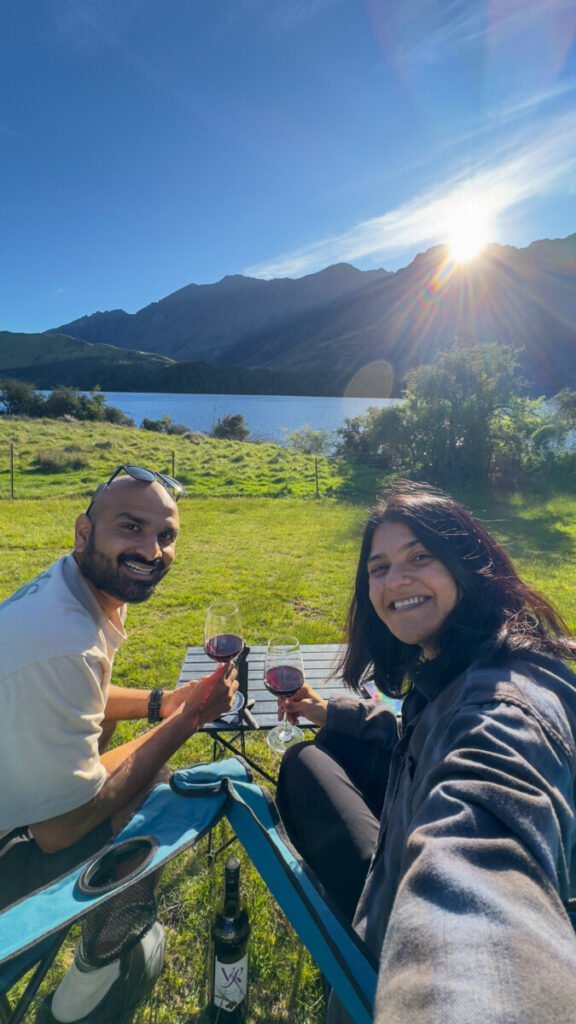 Couple enjoying wine at a picnic table by Moke Lake DOC campsite at sunset with mountains behind them Queenstown New Zealand