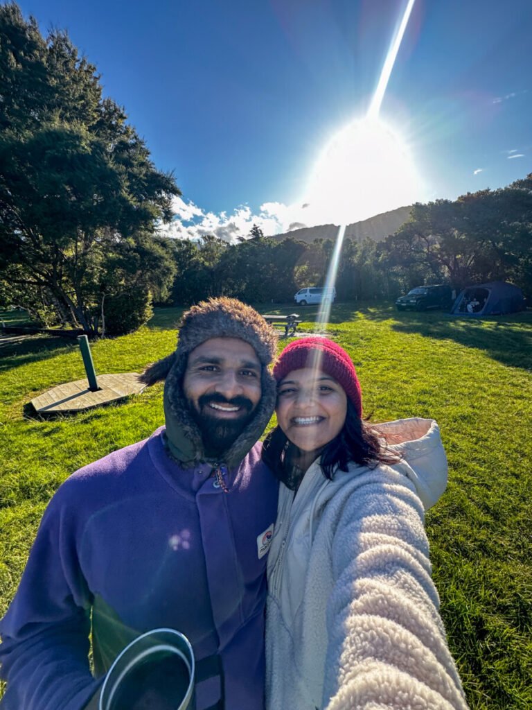 Couple taking a selfie at Momorangi Bay DOC campsite with campervans and forested hills in the background Marlborough New Zealand