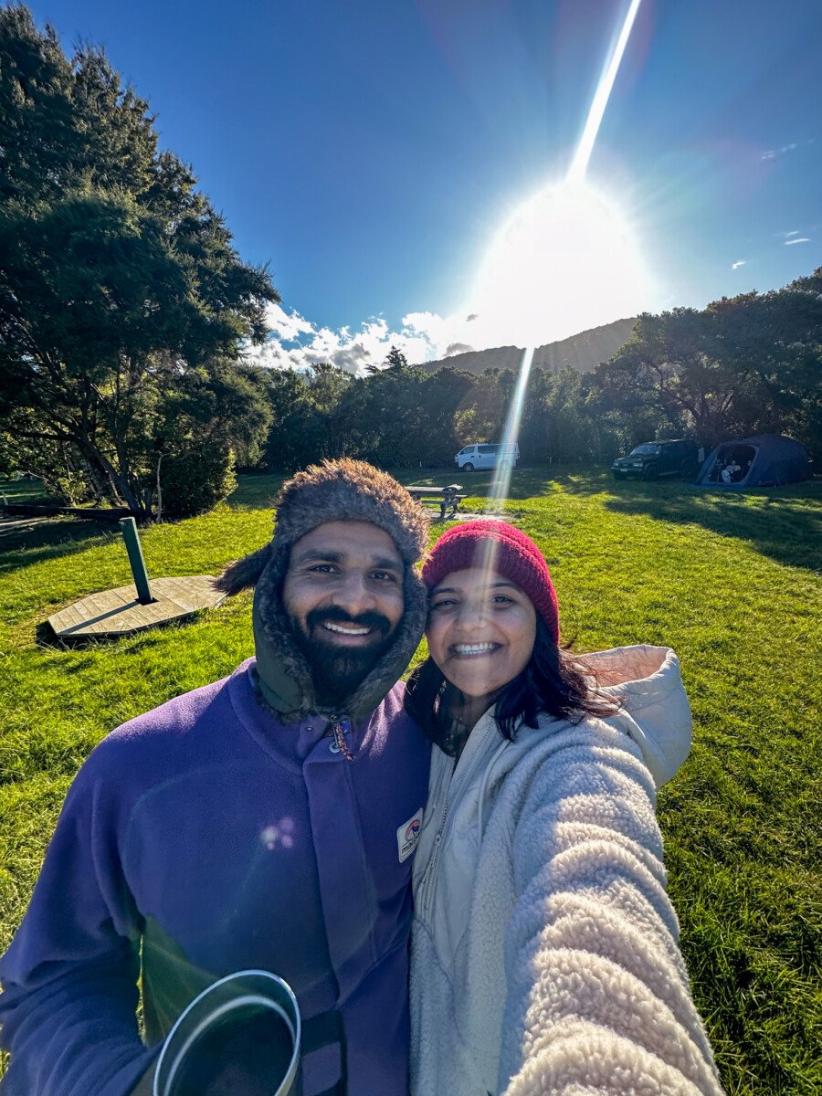 Couple taking a selfie at Momorangi Bay DOC campsite with campervans and forested hills in the background Marlborough New Zealand
