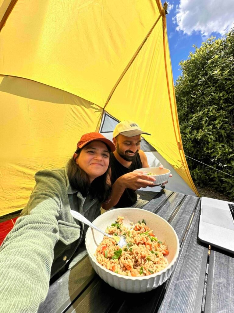 Couple sitting under a yellow camping awning at a picnic table eating a meal from a bowl at their New Zealand campsite