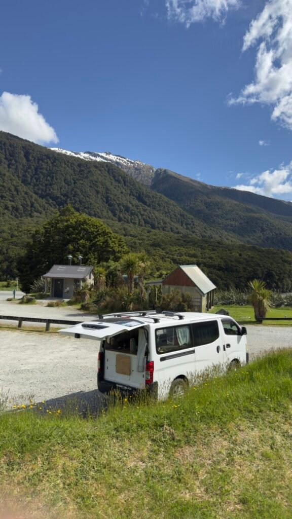 White campervan parked at a DOC campsite with snow-capped mountains and native bush behind it near Lake Hawea in the Queenstown Otago region New Zealand