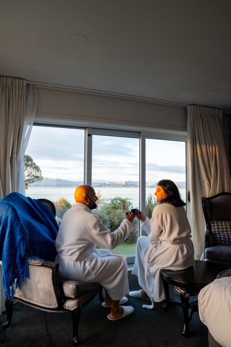Couple in white robes sitting by a large window with Lake Rotorua views at Black Swan Boutique Hotel