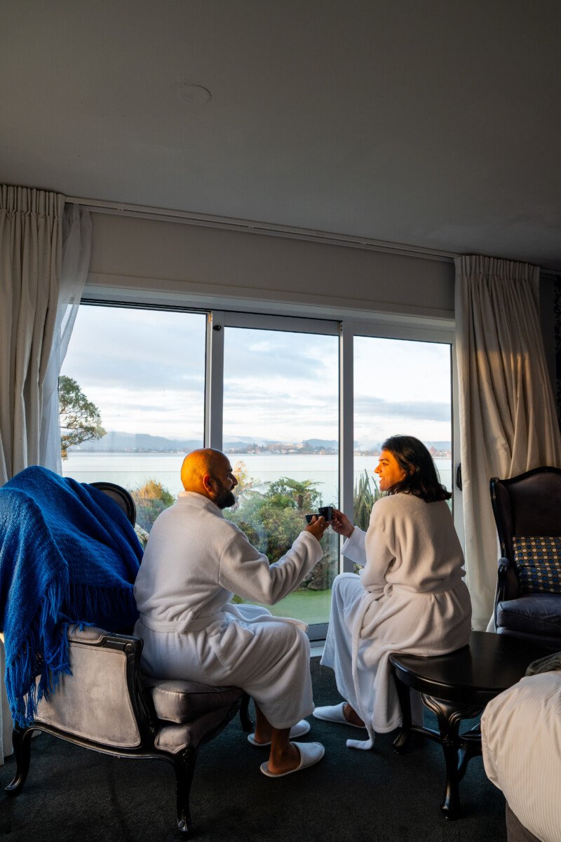 Couple in white robes sitting by a large window with Lake Rotorua views at Black Swan Boutique Hotel