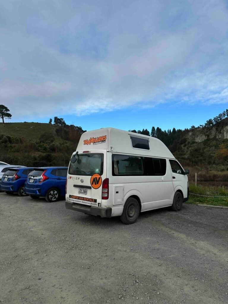 Rear view of a white high-top Travellers campervan parked in a gravel lot with green New Zealand hills behind it