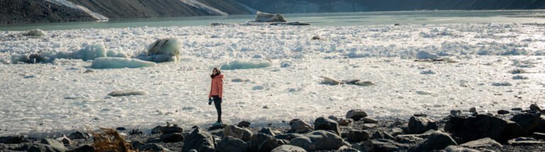 Person in an orange jacket standing on a rocky shore looking out over a glacial landscape toward snow-capped mountains in Aoraki Mount Cook National Park, New Zealand.