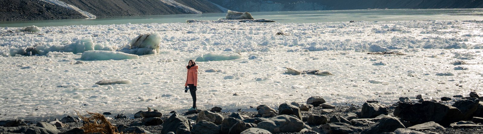 Person in an orange jacket standing on a rocky shore looking out over a glacial landscape toward snow-capped mountains in Aoraki Mount Cook National Park, New Zealand.